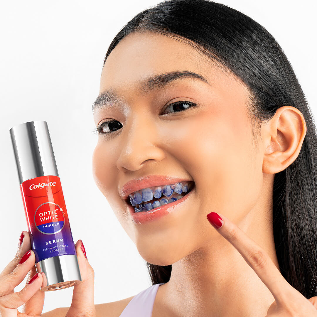 Seorang gadis muda memamerkan botol Serum Optic White Purple dan giginya, yang telah dioleskan serum